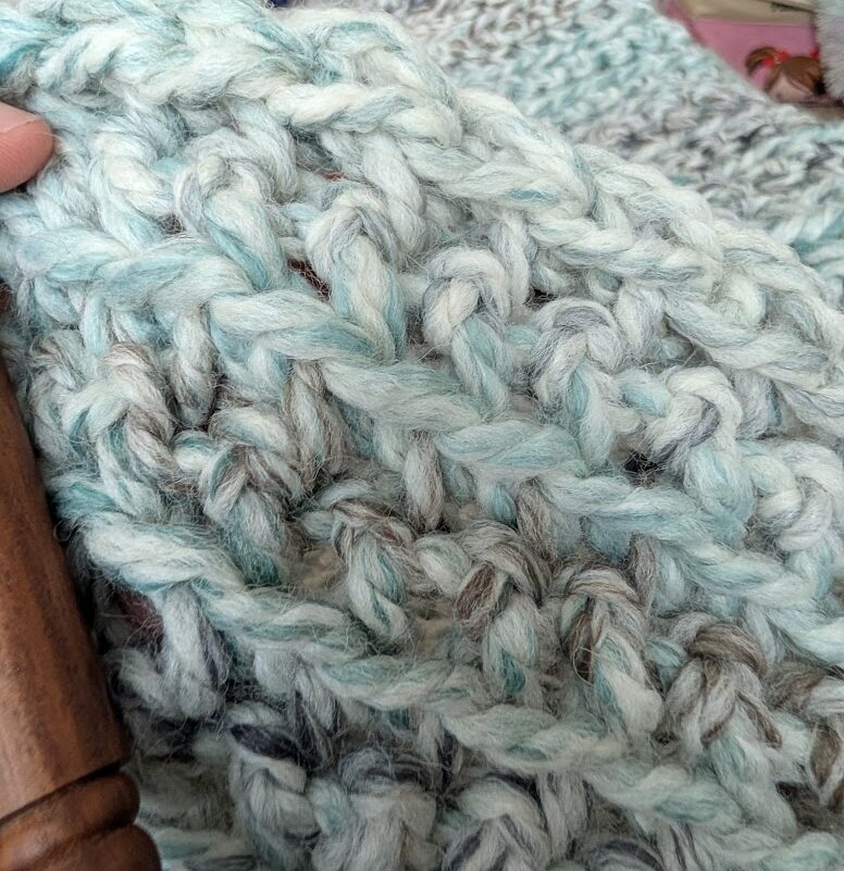 Close-up of a chunky crochet blanket in progress in pale teal, cream, and gray yarn, held at the edge by a thumb.