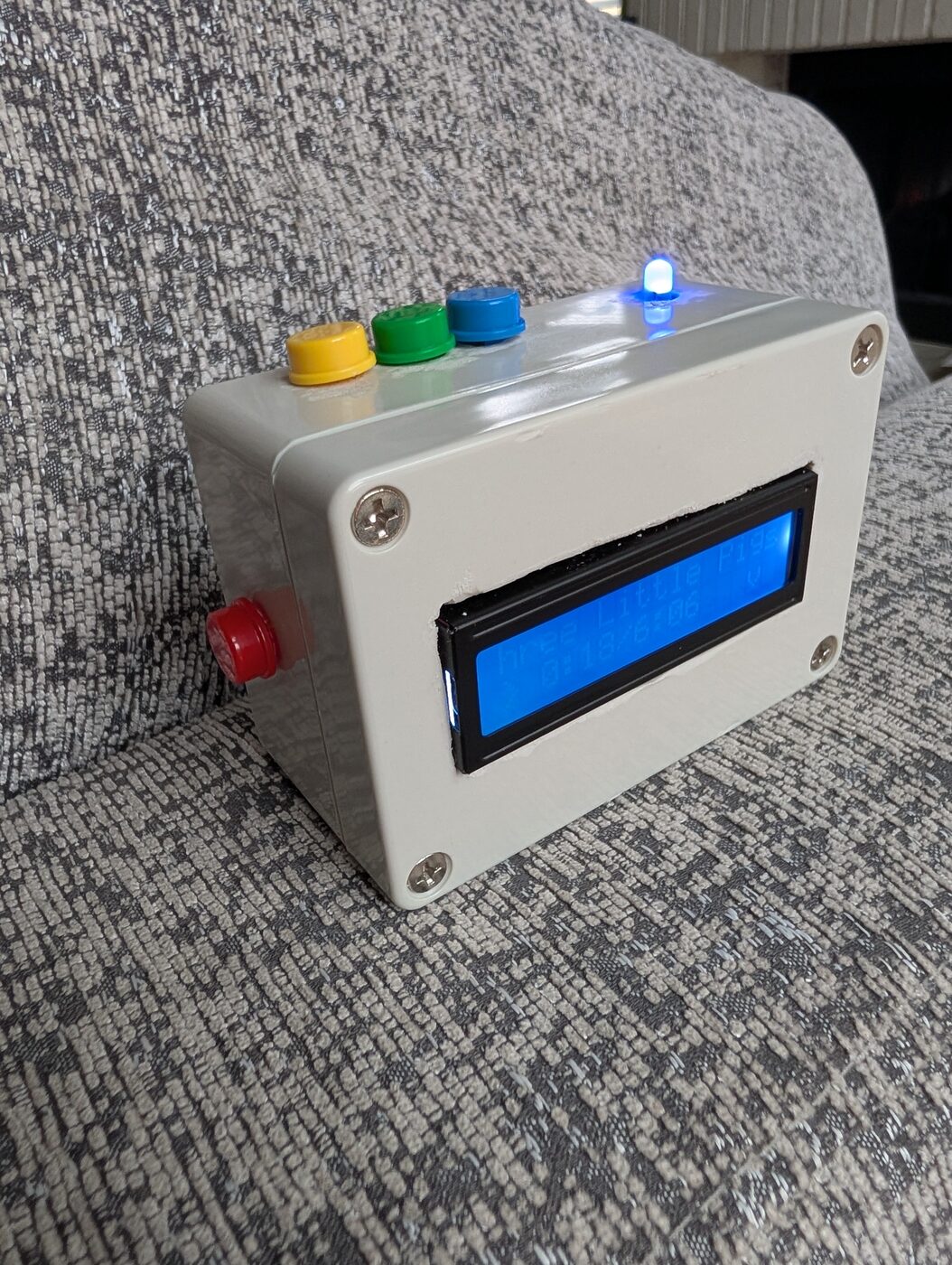 A small white box with three colored buttons on top, a glowing blue LCD on the front, and a status light on.